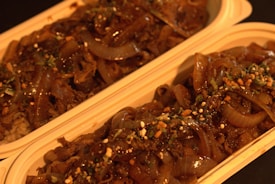 Two rectangular containers are filled with a savory dish consisting of caramelized onions and what appears to be slices of beef or a similar protein, garnished with herbs and small bits of seasoning. The food has a glossy appearance due to the sauce, and the containers are placed side by side.