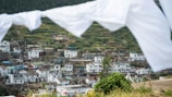A village is nestled in a hilly landscape with terraced fields and whitewashed buildings. The houses are closely packed, featuring both modern and traditional architectural styles. White sheets or flags are hanging and slightly blurred in the foreground, framing the scene. The image conveys a sense of rural life and community.