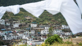A village is nestled in a hilly landscape with terraced fields and whitewashed buildings. The houses are closely packed, featuring both modern and traditional architectural styles. White sheets or flags are hanging and slightly blurred in the foreground, framing the scene. The image conveys a sense of rural life and community.