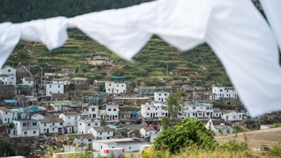 A village is nestled in a hilly landscape with terraced fields and whitewashed buildings. The houses are closely packed, featuring both modern and traditional architectural styles. White sheets or flags are hanging and slightly blurred in the foreground, framing the scene. The image conveys a sense of rural life and community.