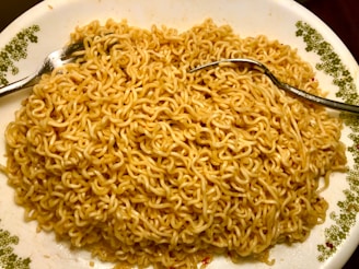A plate filled with cooked instant noodles, with subtle specks of seasoning visible on the noodles. Two silver forks are placed on the dish, one on each side, suggesting the meal is ready to be eaten. The plate has a decorative green floral pattern on the edges.