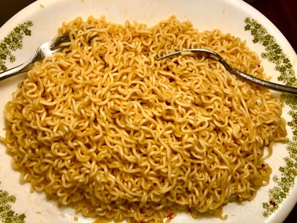 A plate filled with cooked instant noodles, with subtle specks of seasoning visible on the noodles. Two silver forks are placed on the dish, one on each side, suggesting the meal is ready to be eaten. The plate has a decorative green floral pattern on the edges.