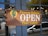 The image features a wooden sign with the words 'Pioneer Saloon OPEN Plate Lunch'. The sign includes a design with a green cactus and a yellow sun against a mountain-like shape. It is affixed to a door with glass panes, through which reflections and a street scene with vehicles can be seen.
