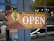 The image features a wooden sign with the words 'Pioneer Saloon OPEN Plate Lunch'. The sign includes a design with a green cactus and a yellow sun against a mountain-like shape. It is affixed to a door with glass panes, through which reflections and a street scene with vehicles can be seen.