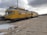 A yellow maintenance train is stationed on a concrete area, with overcast clouds above. The train has multiple sections connected, with windows along the sides, and technical structures on top. Snow patches are visible on the ground.