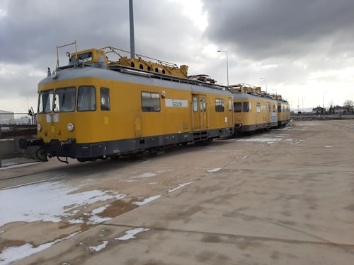 A yellow maintenance train is stationed on a concrete area, with overcast clouds above. The train has multiple sections connected, with windows along the sides, and technical structures on top. Snow patches are visible on the ground.