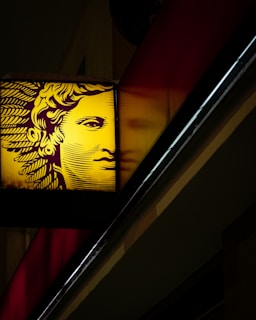 A stylized face, possibly inspired by classical art, is illuminated in yellow against a dark background. The image shows the face on a sign with intricate lines forming hair and features. It is positioned diagonally with a partial reflection beside it.