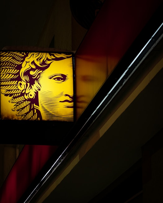 A stylized face, possibly inspired by classical art, is illuminated in yellow against a dark background. The image shows the face on a sign with intricate lines forming hair and features. It is positioned diagonally with a partial reflection beside it.