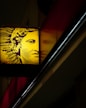 A stylized face, possibly inspired by classical art, is illuminated in yellow against a dark background. The image shows the face on a sign with intricate lines forming hair and features. It is positioned diagonally with a partial reflection beside it.