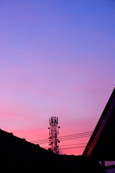 A telecommunication tower stands against a vibrant sunset sky with hues of purple, blue, and pink. The silhouette of rooftops is visible in the foreground.