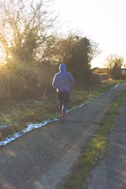 A person wearing a blue hoodie and dark athletic pants is jogging along a paved path. The scene is set during a sunny day with bare trees and some patches of snow or frost on the grass. The sun is low, casting long shadows along the path, and the overall landscape is a mix of greenery and winter elements.
