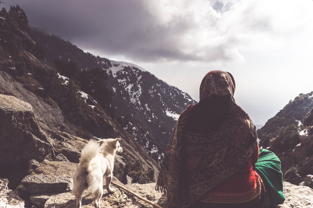 person in black and red jacket sitting on rock near white dog during daytime