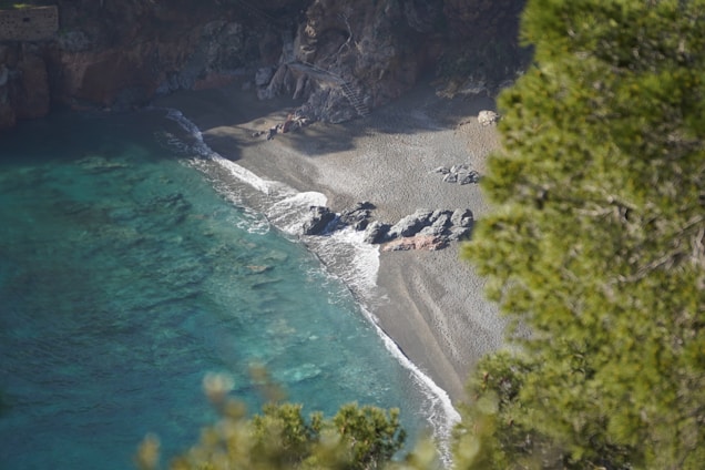 A secluded beach with a mix of pebbles and sand, bordered by clear turquoise water. Rocky formations and green vegetation can be seen in the background. The beach appears tranquil and untouched.