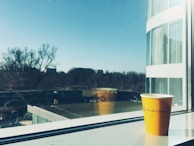 A yellow cup with the Utrecht University logo is placed on a windowsill, with an outside view showing a clear blue sky, distant trees, and modern buildings. The cup has a shadow cast onto the windowsill and there is a reflection of the cup in the window. The scene suggests an early, bright morning.