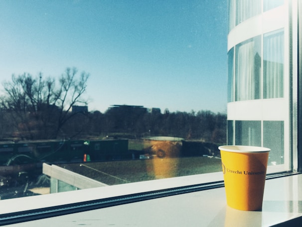 A yellow cup with the Utrecht University logo is placed on a windowsill, with an outside view showing a clear blue sky, distant trees, and modern buildings. The cup has a shadow cast onto the windowsill and there is a reflection of the cup in the window. The scene suggests an early, bright morning.