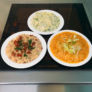 Three plates of creamy dishes are arranged on a smooth, dark surface. The first dish contains a risotto-like meal garnished with chopped herbs and bits of crispy bacon. The second plate holds a green-infused dish with what appears to be small vegetable pieces mixed into a creamy sauce. The third plate features a bright orange dish topped with sliced leeks or onions, suggesting a cheesy or tomato-infused pasta or rice dish.