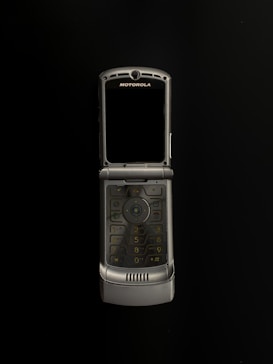 A silver Motorola flip phone is open and centered against a black background. The phone features a numeric keypad and a screen above it, with the brand name visible at the top.