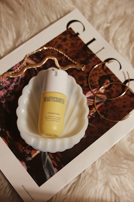A skincare product labeled 'Beauty Counter' placed in a scalloped white dish, sitting on top of a glossy magazine with fashion imagery. Accompanying accessories include a curved gold chain and large hoop earrings, all set against a white, furry background.
