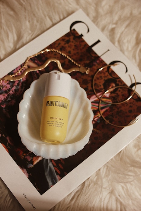 A skincare product labeled 'Beauty Counter' placed in a scalloped white dish, sitting on top of a glossy magazine with fashion imagery. Accompanying accessories include a curved gold chain and large hoop earrings, all set against a white, furry background.