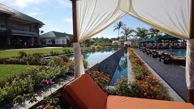 A luxurious outdoor setting featuring a pool surrounded by vibrant orange and yellow flowers. Lounge chairs line the side of the pool, which reflects the blue sky and palm trees. A green-roofed building is visible in the background, while the foreground shows a fabric canopy with a cushioned seat.