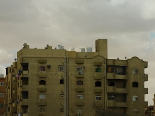 A multi-story residential building with a flat roof, featuring numerous windows and several balconies. The structure appears to be made of concrete with a light brown color. There are water tanks and satellite dishes on the roof, indicating urban living. The sky is overcast with thick gray clouds.