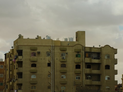 A multi-story residential building with a flat roof, featuring numerous windows and several balconies. The structure appears to be made of concrete with a light brown color. There are water tanks and satellite dishes on the roof, indicating urban living. The sky is overcast with thick gray clouds.