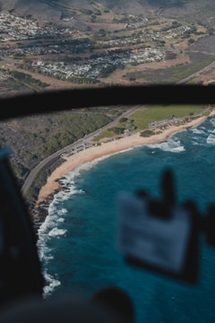 An aerial view of a coastline featuring a sandy beach bordered by turquoise ocean waves. A road runs parallel to the shoreline, with parked cars visible. Inland areas include densely packed buildings, greenery, and hills in the background.