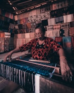 An older woman with glasses is weaving textiles on a traditional loom. She is surrounded by a variety of colorful fabrics displayed on the walls behind her, creating a visually intricate background. The lighting in the area is warm, highlighting the rich textures and colors of the fabrics. The woman is intensely focused on her work.