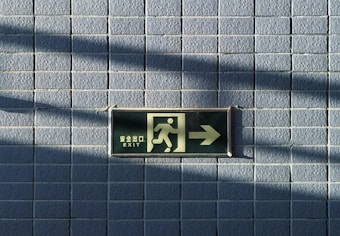 A green exit sign is mounted on a tiled wall, featuring a running figure and an arrow pointing to the right. The sign includes text in both English and another language. Shadows cast on the wall create a pattern, adding depth to the image.