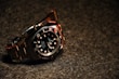 A luxury wristwatch with a black dial and steel bracelet is prominently displayed. The watch features a rotating bezel with large, luminous markers and hands.