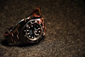 A luxury wristwatch with a black dial and steel bracelet is prominently displayed. The watch features a rotating bezel with large, luminous markers and hands.