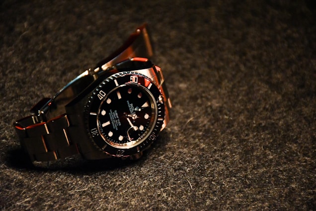 A luxury wristwatch with a black dial and steel bracelet is prominently displayed. The watch features a rotating bezel with large, luminous markers and hands.