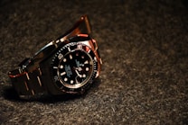 A luxury wristwatch with a black dial and steel bracelet is prominently displayed. The watch features a rotating bezel with large, luminous markers and hands.