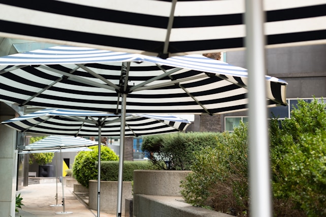 A row of large striped umbrellas lines an outdoor area, shading a walkway next to neatly trimmed bushes and planters. The surrounding environment includes concrete structures and a modern building facade.