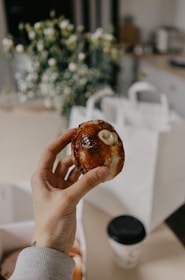 A hand holds a glazed pastry with a white filling at the top. The background includes a blurred arrangement of white flowers and white paper bags, with a takeaway coffee cup visible on the table.