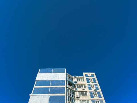 A multi-story building with a modern architectural design features large, reflective windows and solar panels. The structure occupies the lower portion of the composition, while the upper area is dominated by a vast, clear blue sky.