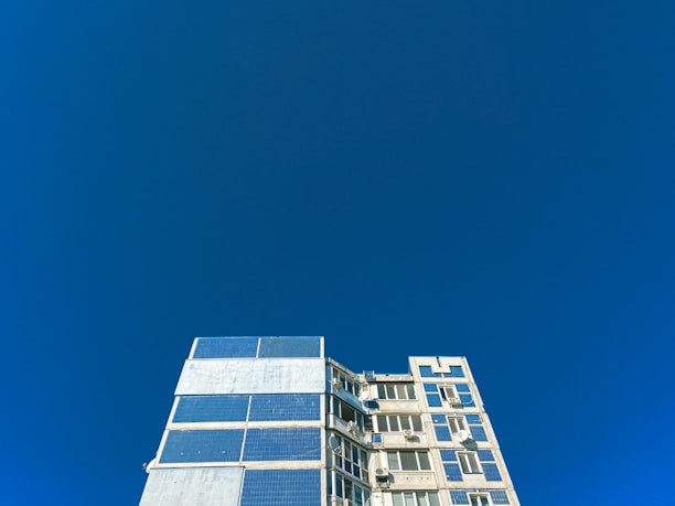 A multi-story building with a modern architectural design features large, reflective windows and solar panels. The structure occupies the lower portion of the composition, while the upper area is dominated by a vast, clear blue sky.