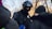 A police officer wearing a helmet and protective gear appears to be in a conversation with individuals dressed in winter clothing. The scene takes place outdoors, indicated by bare trees in the background, suggesting a cold environment.