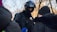 A police officer wearing a helmet and protective gear appears to be in a conversation with individuals dressed in winter clothing. The scene takes place outdoors, indicated by bare trees in the background, suggesting a cold environment.