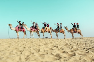 A line of camels carrying people dressed in colorful attire, raising their arms in celebration, traverse a sandy desert landscape beneath a vast blue sky.