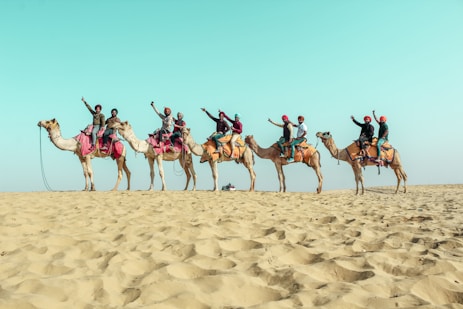 A line of camels carrying people dressed in colorful attire, raising their arms in celebration, traverse a sandy desert landscape beneath a vast blue sky.