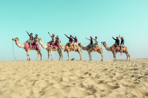 A line of camels carrying people dressed in colorful attire, raising their arms in celebration, traverse a sandy desert landscape beneath a vast blue sky.