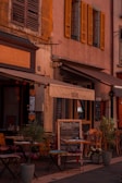 A charming outdoor café with small tables and chairs set up on a cobblestone street, surrounded by potted plants. The café has a rustic appearance, featuring weathered shutters and a warm color palette. A menu board is displayed outside.