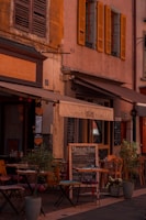 A charming outdoor café with small tables and chairs set up on a cobblestone street, surrounded by potted plants. The café has a rustic appearance, featuring weathered shutters and a warm color palette. A menu board is displayed outside.