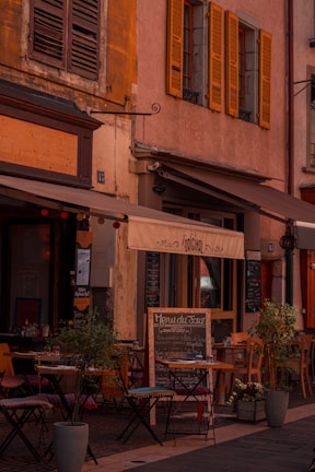 A charming outdoor café with small tables and chairs set up on a cobblestone street, surrounded by potted plants. The café has a rustic appearance, featuring weathered shutters and a warm color palette. A menu board is displayed outside.