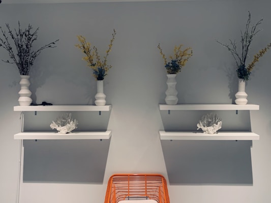 Two white shelves are mounted on a gray wall, each holding a white vase with decorative branches and yellow flowers. Below each shelf is another shelf with a white coral sculpture. An orange wire basket is partially visible at the bottom of the image.