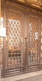 A large wrought iron gate with intricate floral and geometric patterns, featuring golden accents. The structure is set against a solid backdrop and has a keypad entry system on the left side.