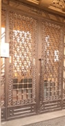A large wrought iron gate with intricate floral and geometric patterns, featuring golden accents. The structure is set against a solid backdrop and has a keypad entry system on the left side.