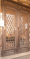 A large wrought iron gate with intricate floral and geometric patterns, featuring golden accents. The structure is set against a solid backdrop and has a keypad entry system on the left side.