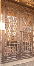 A large wrought iron gate with intricate floral and geometric patterns, featuring golden accents. The structure is set against a solid backdrop and has a keypad entry system on the left side.